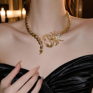 May include: A gold-toned metal choker necklace featuring a segmented, curved body that wraps around the neck. The design includes a detailed scorpion figure at the centre, with the scorpion's head and thorax embellished with clear, sparkling crystals.