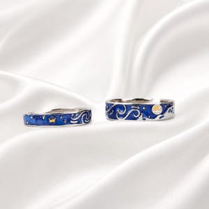 Puede incluir: Dos anillos plateados con una incrustación de esmalte azul profundo con patrones de remolinos, pequeñas estrellas doradas y una luna creciente. Las bandas están abiertas en la parte posterior para ajustarse y tienen un acabado metálico pulido.