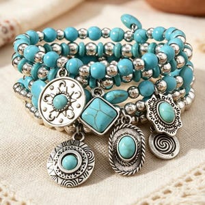 Op de afbeelding: Een meerrijige wikkelarmband met turkooiskleurige kralen en metallic zilverkleurige kralen. De armband bevat diverse hangende bedels, waaronder cirkelvormige en ruitvormige hangers met ingelegde turkooiskleurige stenen en gegraveerde patronen.