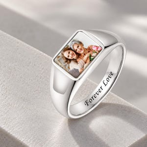 Puede incluir: Un anillo de sello en tono plateado con una fotografía rectangular incrustada de dos personas abrazadas sosteniendo flores rosas. El lateral de la banda de metal pulido tiene grabado el texto Forever Love en una fuente cursiva clara.
