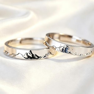 May include: Two silver-toned metal rings featuring engraved designs. One ring displays a mountain range with dark accents, while the other ring features a stylised ocean wave and smaller mountain peaks, both with polished, reflective surfaces.