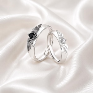 May include: A pair of silver-toned rings featuring distinct designs. One ring displays a black square-cut stone set within bat-like wings, while the other ring features a clear round-cut stone accented by delicate, stylized angel-like wings.