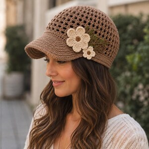 May include: A brown crocheted baseball cap featuring an open-weave grid pattern on the crown and a solid brim. The cap is decorated with two cream-coloured crocheted flowers with brown centres and a cluster of dark green crocheted leaves.