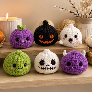 May include: Six small, round, crocheted plush figures arranged in two rows. The top row features a purple pumpkin with a green stem, a black jack-o'-lantern, and a white ghost. The bottom row includes a green creature, a white skull, and a purple monster.