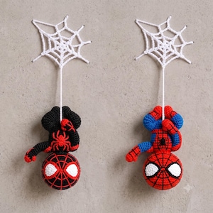 Puede incluir: Dos figuras de ganchillo colgando de telarañas de hilo blanco. Una figura es negra con un emblema de araña rojo, la otra es roja y azul con un emblema negro. Ambas tienen grandes ojos blancos y cabezas redondas.