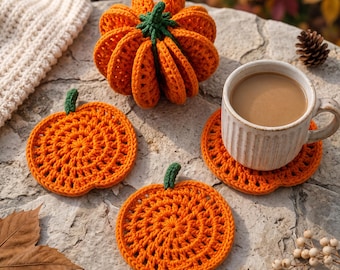 May include: A three-dimensional crocheted orange pumpkin with a green stem, accompanied by two flat, circular crocheted pumpkin-shaped coasters in orange with green stems, and a white ceramic mug filled with a light brown beverage.