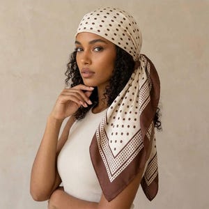Puede incluir: Un pañuelo cuadrado con base color crema, estampado de lunares marrones y un borde ancho marrón con detalles de puntos más pequeños. El pañuelo está atado alrededor de la cabeza, cae por el lado y se lleva con una camiseta sin mangas color crema.