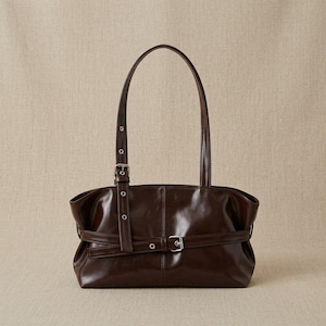 Peut inclure: Un sac à épaule marron foncé à la finition brillante, doté d'une ceinture horizontale sur le devant avec une boucle en métal argenté et d'une bandoulière réglable assortie avec des œillets argentés et une fermeture à boucle.