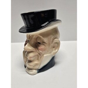 May include: A ceramic character jug shaped like a person's head wearing a black top hat. The figure has a beige complexion, defined facial features, and a dark collar. The glossy finish highlights the sculpted details of the face and the hat.