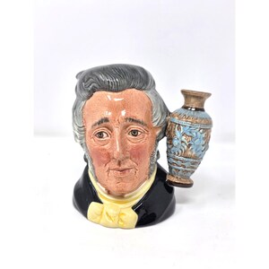 May include: A ceramic character jug depicting a person with gray hair, wearing a black coat and a yellow cravat. The handle of the jug is shaped like a small, light blue and brown decorative vase with intricate floral relief patterns on its surface.