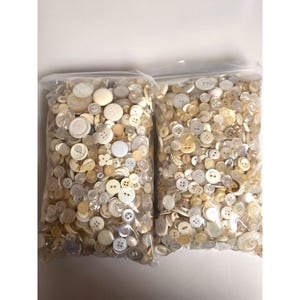 May include: Two clear plastic bags filled with a large assortment of loose buttons in various shades of white, cream, and beige. The buttons vary in size, shape, and number of holes, featuring both flat and textured surfaces throughout the collection.