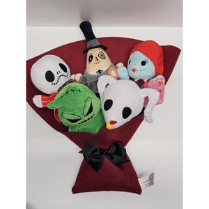 May include: A plush bouquet featuring five characters from The Nightmare Before Christmas, including Jack Skellington, Sally, Zero, Oogie Boogie, and the Mayor, wrapped in dark red felt and tied with a black ribbon bow at the base.