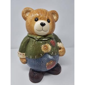 May include: A ceramic teddy bear figurine featuring a brown bear wearing a textured green sweater and blue pants. The clothing has visible stitched patches in tan and red with button details. The bear has a smiling face and dark brown shoes.