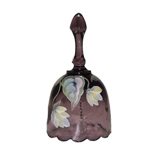May include: A translucent purple glass bell featuring a hand-painted floral design with light green and white petals. The bell has a scalloped bottom edge and a slender, tapered handle at the top, with a thin metal chain visible inside the glass body.