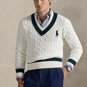 May include: A white cable-knit V-neck sweater featuring dark green and navy blue striped trim at the neckline, cuffs, and hem. A navy blue embroidered polo player logo is on the left chest. The sweater is worn over a blue and white striped shirt.