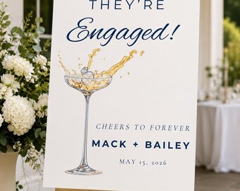 Könnte beinhalten: Ein weißes rechteckiges Hochzeitsschild auf einer goldenen Staffelei mit dem Text THEY'RE Engaged! CHEERS TO FOREVER MACK + BAILEY MAY 15, 2026. Das Design enthält eine Aquarellillustration eines Champagnerglases mit einem Diamantring.