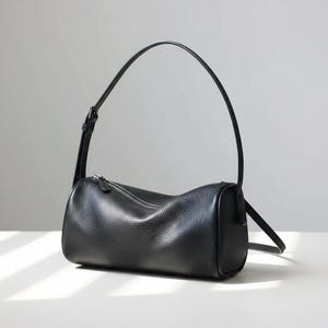 May include: A black cylindrical handbag with a textured leather-like finish, featuring a silver-toned metal zipper closure along the top and a single adjustable shoulder strap with a small metal buckle for length customization.