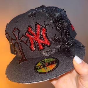 Pode incluir: Um boné de beisebol preto com um logótipo dos New York Yankees e um design de cruz adornados com cristais vermelhos. O boné é decorado com rosas de tecido preto e missangas na lateral. Um autocolante circular na pala mostra New Era 59FIFTY 7 1/4 (57,7 cm).