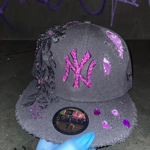 May include: A charcoal grey New Era 59FIFTY baseball cap with a size 7 3/8 sticker on the brim. The cap features a pink rhinestone NY logo, black floral lace appliqué, scattered pink rhinestones, and distressed fabric patches with purple fabric.
