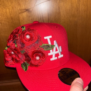 Peut inclure: Une casquette de baseball rouge vif ornée d'un logo LA blanc brodé sur le devant et d'un groupe de roses en tissu rouge avec de petites perles blanches au centre sur le côté. Un autocollant noir 59FIFTY est apposé sur la visière plate.