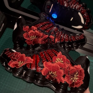 Könnte beinhalten: Ein Paar schwarze Sneaker mit einem glänzenden, roten Krokodil-Texturmuster an den Seiten und großen, detaillierten roten Blumenstickereien. Die Schuhe haben schwarze Sohlen mit sichtbaren Dämpfungssäulen und ein schwarzes Nike-Logo.