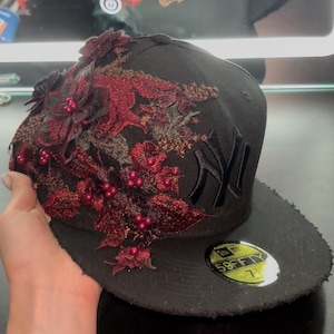 Può includere: Un cappellino da baseball nero con un logo dei New York Yankees ricamato in nero. Il lato è decorato con intricati inserti in pizzo floreale rosso scuro e piccole perline rosse lucide. Un adesivo circolare 59FIFTY si trova sulla visiera piatta.