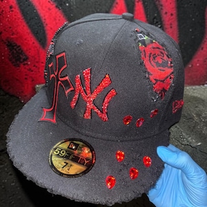 Pode incluir: Um boné de beisebol preto New Era 59FIFTY com um adesivo de tamanho 7 1/4 na aba. O boné apresenta um logotipo NY de strass vermelho, um emblema de cruz vermelha, um rasgo com padrão de rosa vermelha e strass vermelhos em forma de gota.