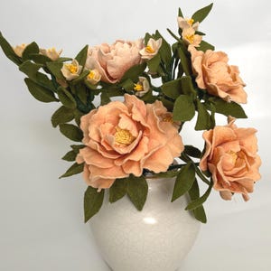 May include: A bouquet of peach-colored felt flowers with yellow centers and dark green felt leaves arranged in a white ceramic vase. The vase features a subtle crackle glaze pattern across its rounded surface, holding the floral arrangement.
