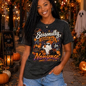 May include: A dark heather gray short-sleeve t-shirt featuring a graphic print of a ghost wearing sunglasses and an orange beanie, holding a mug. The text reads Seasonally UNAVAILABLE for Nonsense, surrounded by pumpkins, a lantern, and autumn leaves.