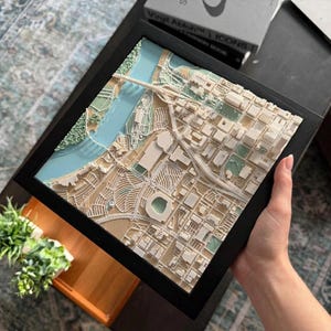 Puede incluir: Un mapa topográfico 3D enmarcado de forma cuadrada que muestra un plano de ciudad con edificios beige, un río azul claro y zonas verdes. El modelo detallado incluye un puente sobre el agua y un estadio dentro de la cuadrícula.