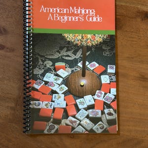 Könnte beinhalten: Ein spiralgebundenes Lehrbuch mit dem Titel American Mahjong: A Beginner's Guide. Das Cover zeigt eine orangefarbene Kopfzeile, einen grünen Streifen und ein Foto einer Holzlampe, umgeben von weißen und orangefarbenen Mahjong-Spielsteinen.