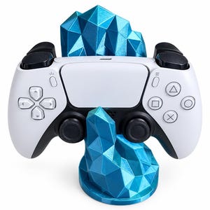 May include: A white and black wireless game controller resting on a bright blue, geometric, crystal-shaped stand. The stand features a faceted, angular design that supports the controller in an upright position, showcasing the buttons and joysticks.