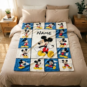 May include: A soft rectangular blanket featuring a grid of blue squares, each displaying a different illustration of Mickey Mouse. The centre panel is white with a large Mickey Mouse graphic and the word NAME printed in black text above it.