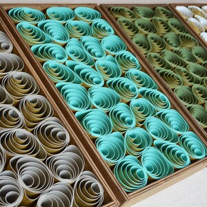 May include: Four rectangular wooden frames containing rows of tightly coiled paper strips. The first frame features gray and yellow coils, the second turquoise, the third olive green, and the fourth white and cream, creating a textured, layered effect.