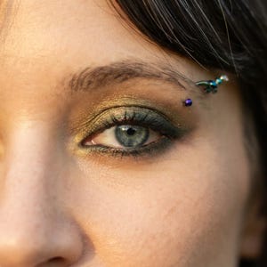 Può includere: Un primo piano di un occhio blu-grigio con ombretto oro metallizzato e verde scuro applicato sulla palpebra. Un piccolo piercing al sopracciglio curvo con una perlina viola scuro e un accento blu-verde metallizzato è visibile sull'arcata.