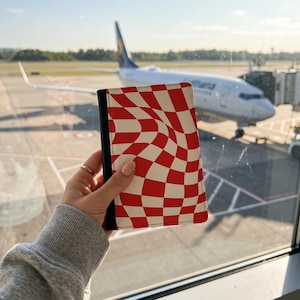 May include: A rectangular passport holder featuring a red and white wavy checkerboard pattern on the front cover, with a solid black spine detail. The cover displays a distorted grid design with alternating red and white squares.