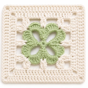 May include: A square crochet motif featuring a central four-leaf clover design in light green yarn, surrounded by a cream-coloured border with an openwork pattern. The piece is constructed with visible yarn stitches forming a geometric square shape.