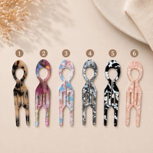 Puede incluir: Seis horquillas para el pelo dispuestas en fila, numeradas del uno al seis. Cada horquilla tiene una parte superior circular y dos puntas con aberturas rectangulares. Los diseños incluyen carey marrón, morado y azul, rosa y azul, gris, negro y blanco, y rosa.