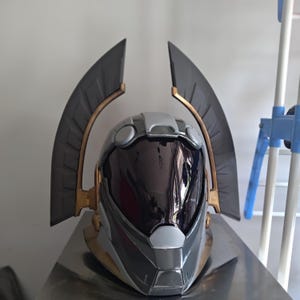 Puede incluir: Un casco futurista con una visera oscura y reflectante, una carcasa segmentada en gris claro y gris oscuro, y dos grandes crestas curvas en forma de ala que se extienden hacia arriba, con detalles en dorado metálico en la base y bordes.