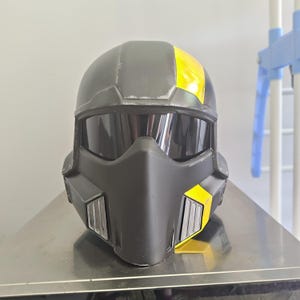 Könnte beinhalten: Ein mattschwarzer Vollvisierhelm mit einem dunkel getönten Visier, einem spitzen Kinnschutz und einem auffälligen, leuchtend gelben Streifen, der vertikal an der rechten Seite verläuft. Der Helm hat silberfarbene Lüftungsschlitze vorne.