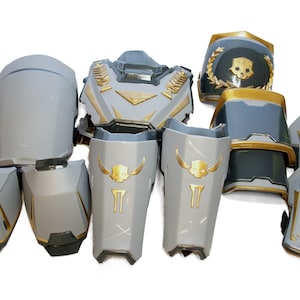 Puede incluir: Un conjunto de piezas de armadura grises y doradas que incluye una placa pectoral, hombreras y grebas. La armadura presenta emblemas de calaveras doradas, coronas de laurel y detalles geométricos, con un acabado liso en todas las piezas.