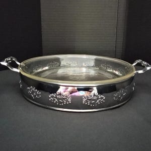 May include: A round glass casserole dish set inside a polished silver-tone metal frame. The metal frame features decorative cut-out patterns along the sides and two side handles. The clear glass insert has a smooth rim and base.