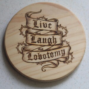 Op de afbeelding: Een ronde onderzetter van lichtgekleurd hout met een brandmerkontwerp van een lint in drie lagen. De tekst "Live Laugh Lobotomy" staat in een zwart gotisch lettertype op het lint, omringd door een gedetailleerde illustratie van doornen.