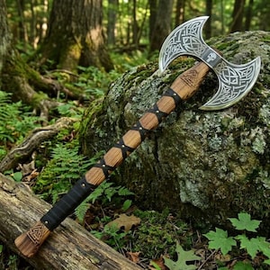 May include: A double-bitted axe featuring a silver-toned metal head with intricate Celtic knotwork engravings and a small skull emblem. The handle is crafted from light brown wood, wrapped with black leather cord in a crisscross pattern.