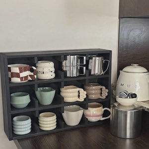 Może przedstawiać: Czarna drewniana p&oacute;łka z przegr&oacute;dkami zawierająca dwanaście ceramicznych kubk&oacute;w i filiżanek o r&oacute;żnych kształtach, kolorach i fakturach, w tym wzory w kratkę, nakrapiane, żebrowane i gładkie, oraz dwa dzbanki ze stali nierdzewnej.