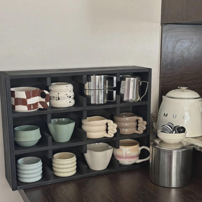 Może przedstawiać: Czarna drewniana p&oacute;łka z przegr&oacute;dkami zawierająca dwanaście ceramicznych kubk&oacute;w i filiżanek o r&oacute;żnych kształtach, kolorach i fakturach, w tym wzory w kratkę, nakrapiane, żebrowane i gładkie, oraz dwa dzbanki ze stali nierdzewnej.