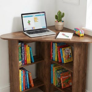 May include: A dark wood corner desk featuring two open shelving units filled with colorful children's books. On the desktop sits an open laptop, a small potted cactus, a spiral-bound notebook with a pen, and a small patterned pencil case.