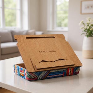 May include: A wooden book stand with a rectangular base featuring a colorful, multi-patterned embroidered fabric border. The light brown wooden surface has a horizontal slot and a central ledge, with the name CAROL SMITH engraved in the center.