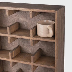 Może przedstawiać: Ciemnobrązowa drewniana p&oacute;łka ścienna z wieloma prostokątnymi przegr&oacute;dkami ułożonymi w kratkę. Pojedynczy jasnobeżowy ceramiczny kubek z zaokrąglonym uchem znajduje się w jednej z g&oacute;rnych sekcji p&oacute;łki, spoczywając na drewnianej powierzchni.