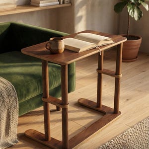 Puede incluir: Una mesa auxiliar rectangular de madera oscura con base en forma de C y esquinas redondeadas. La mesa cuenta con dos soportes cilíndricos verticales conectados por un refuerzo horizontal. Un libro abierto y una taza de cerámica marrón.